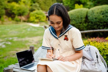 Young smiling asian woman wearing dress writing down notes while sitting on bench in green park