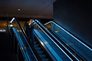Young smiling asian woman wearing suit using mobile phone while standing on escalator indoors