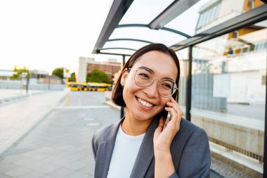 Young smiling asian woman in glasses talking on mobile phone while standing at bus station