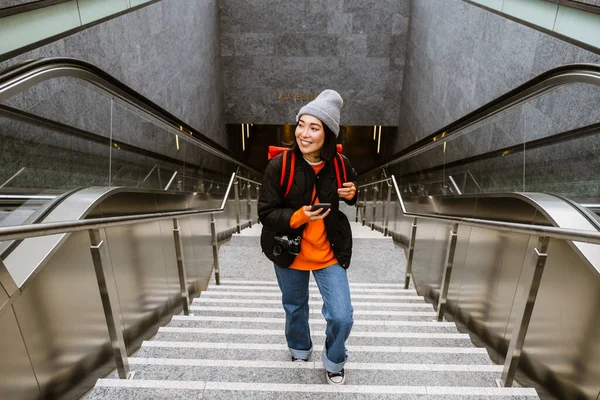 階段を登りながら屋外でスマートフォンを使用して暖かい服で美しい若い笑顔アジアの女性 — ストック写真