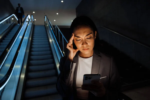 年轻的不高兴的亚洲女人穿着西服 一边皱着眉头一边用智能手机站在室内扶梯上 — 图库照片