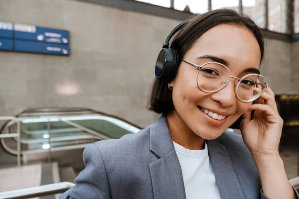 ヘッドフォンで若いアジアの女性は笑顔とエスカレーターの近くに屋外に立っている間カメラを見て — ストック写真