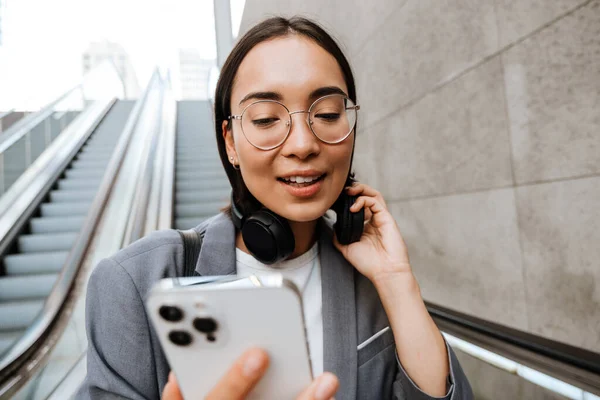 Gözlüklü Akıllı Telefon Kullanan Yürüyen Merdivende Dururken Kulaklık Takan Asyalı — Stok fotoğraf