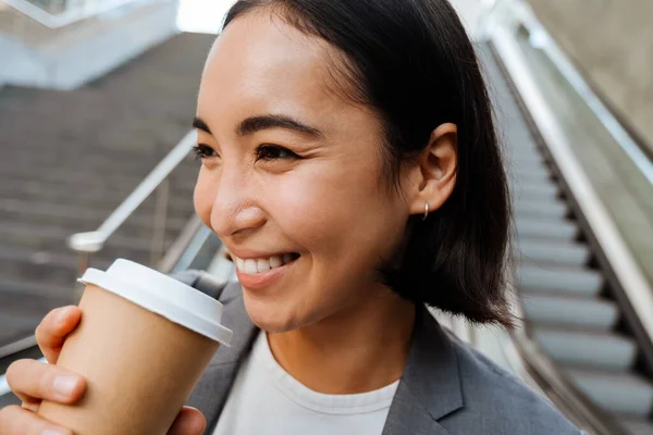 快乐的亚洲女人穿着西服 站在自动扶梯前喝咖啡 — 图库照片