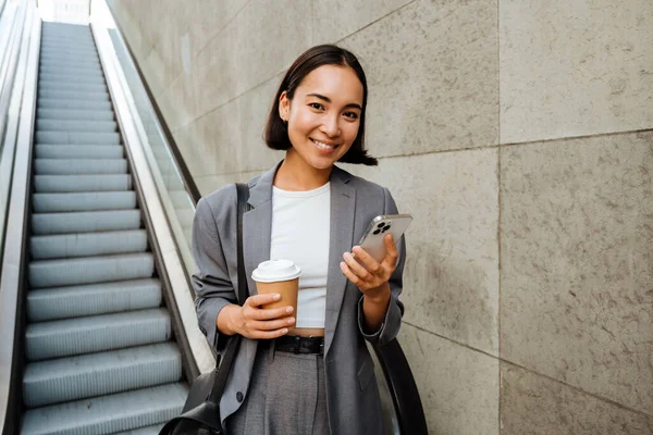 Young Smiling Asian Woman Holding Coffee Using Mobile Phone While — Zdjęcie stockowe