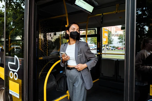 Młoda Azjatka Masce Medycznej Smartfonem Ręku Spoglądającym Bok Stojąc Autobusie — Zdjęcie stockowe