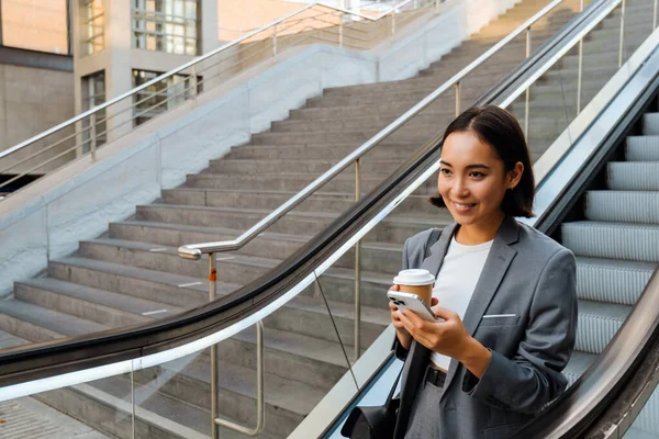 Young Asian Woman Using Cellphone Holding Coffee While Going Escalator — Stockfoto