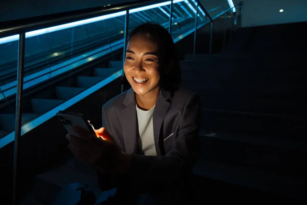 エスカレーターの横の階段に座っている間 携帯電話を使用して若い陽気なアジアの女性 — ストック写真