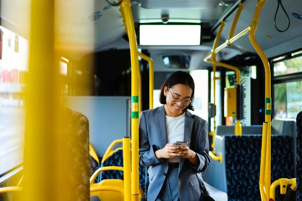 Young asian woman in glasses smiling and using smart phone while standing in a bus