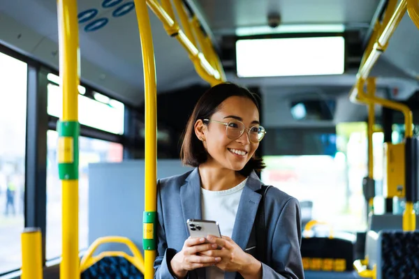 Young asian woman in glasses smiling and using cellphone while standing in a bus
