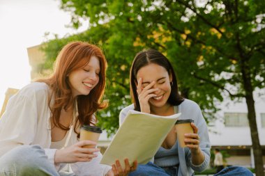 Two young beautiful smiling happy girls with closed eyes sitting in sunny park with coffee and documents