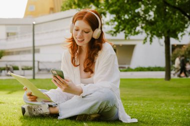 Young bautiful smiling redhead girl in headphones with phone and documents in her hands sitting in lotus pose on the grass