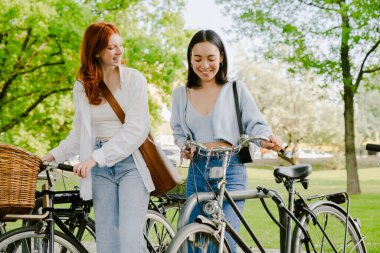 Two young beautiful smiling happy girls walking with bicycles in park