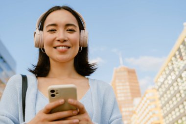 Portrait of young smiling beautiful asian girl in wireless headphones holding phone and looking at camera , while standing in the city on the street
