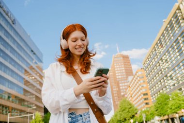 Young beautiful smiling redhead girl in headphones holding her phone both hands and looking on it, while standing in the city on the street