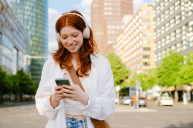 Young beautiful smiling redhead girl in headphones holding her phone both hands and looking on it, while standing in the city on the street