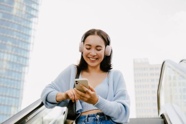 Portrait of young beautiful asian smiling happy girl in headphones using her phone while standing on escalator in the city outdoors