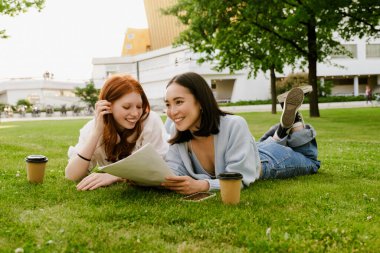 Young multinational women drinking coffee while doing homework in park