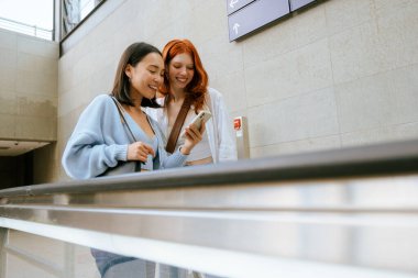 Young two multinational women using mobile phone while standing on escalator at subway station