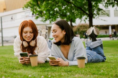 Young multinational women drinking coffee and using cellphone in park