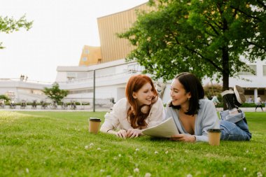 Young multinational women drinking coffee while doing homework in park