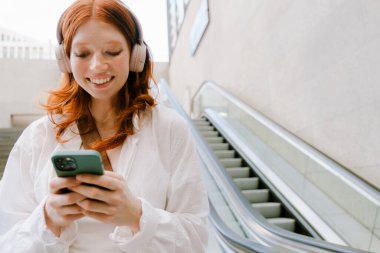 Young white woman wearing headphones using mobile phone while standing on escalator
