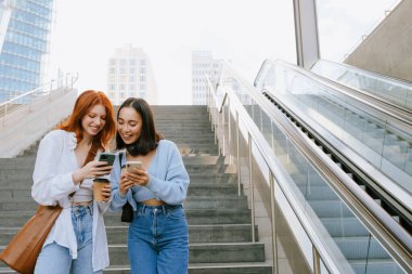 Young multinational women smiling and using cellphones while standing on city stairs