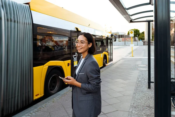 Young asian smiling woman using mobile phone while standing at bus station
