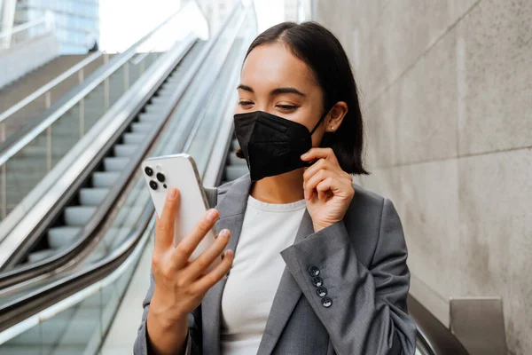 若いですアジアの女性で医療用マスク見ます携帯電話の画面上に立ちながらエスカレーター — ストック写真