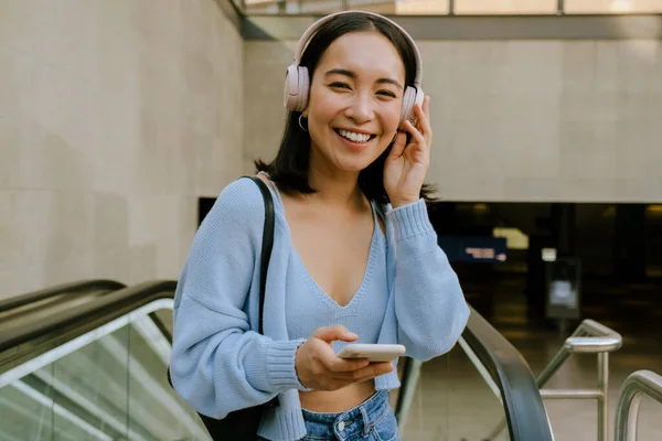 地下鉄駅のエスカレーターに立ちながらヘッドフォンをして携帯電話を使う若いアジア人女性 — ストック写真