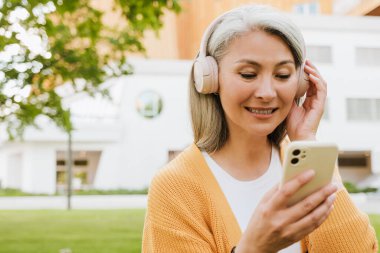 Grey asian woman in headphones laughing and using cellphone while standing in park