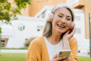 Grey asian woman in headphones laughing and using cellphone while standing in park
