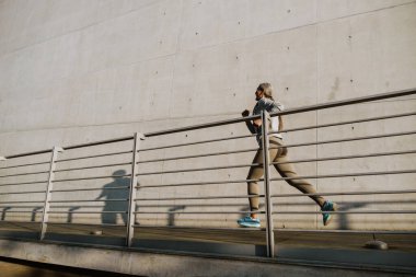 Asian mature woman in tracksuit running by concrete wall during workout outdoors