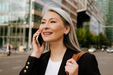 Grey asian woman laughing and talking on cellphone at city street