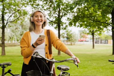 Grey asian woman laughing and using cellphone while standing by her bicycle in park