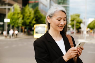 Grey asian woman smiling and using mobile phone at city street