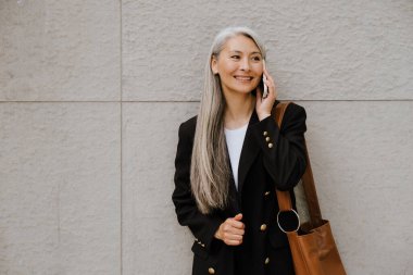 Grey asian woman smiling and talking on cellphone while standing by wall outdoors