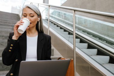Grey asian woman drinking coffee and working with laptop on stair indoors