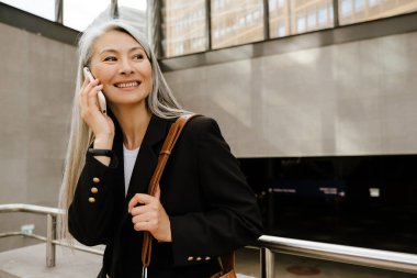 Grey asian woman smiling and talking on cellphone while standing indoors