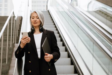 Mature asian woman using mobile phone while standing on escalator indoors