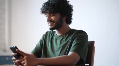 Smiling curly haired Indian man texting on mobile while sitting at home