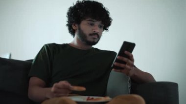 Serious curly haired Indian man texting on mobile and eating pizza while sitting on the couch at home