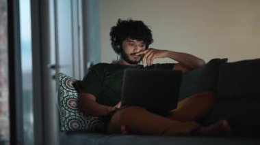 Happy curly haired Indian man working on laptop in headphones on the sofa at home