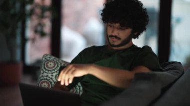 Cheerful curly haired Indian man working on laptop on the sofa at home