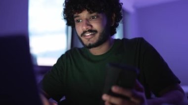 Brunet happy curly haired Indian man typing on laptop at a table at home