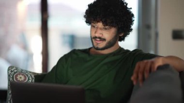 Happy curly haired Indian man working on laptop and talking by video call on the sofa at home