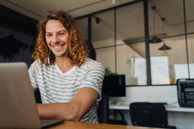 Young curly man using laptop and earphones while sitting by desk in office