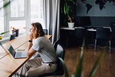 Young curly man smiling and using laptop while sitting by desk in office