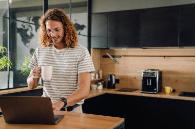 Young curly man drinking coffee by table while working on laptop in office kitchen