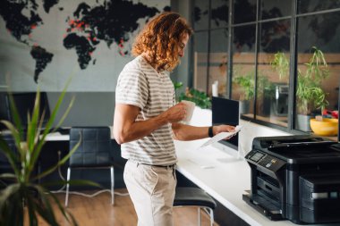 Young curly man drinking coffee and working with copy machine in office